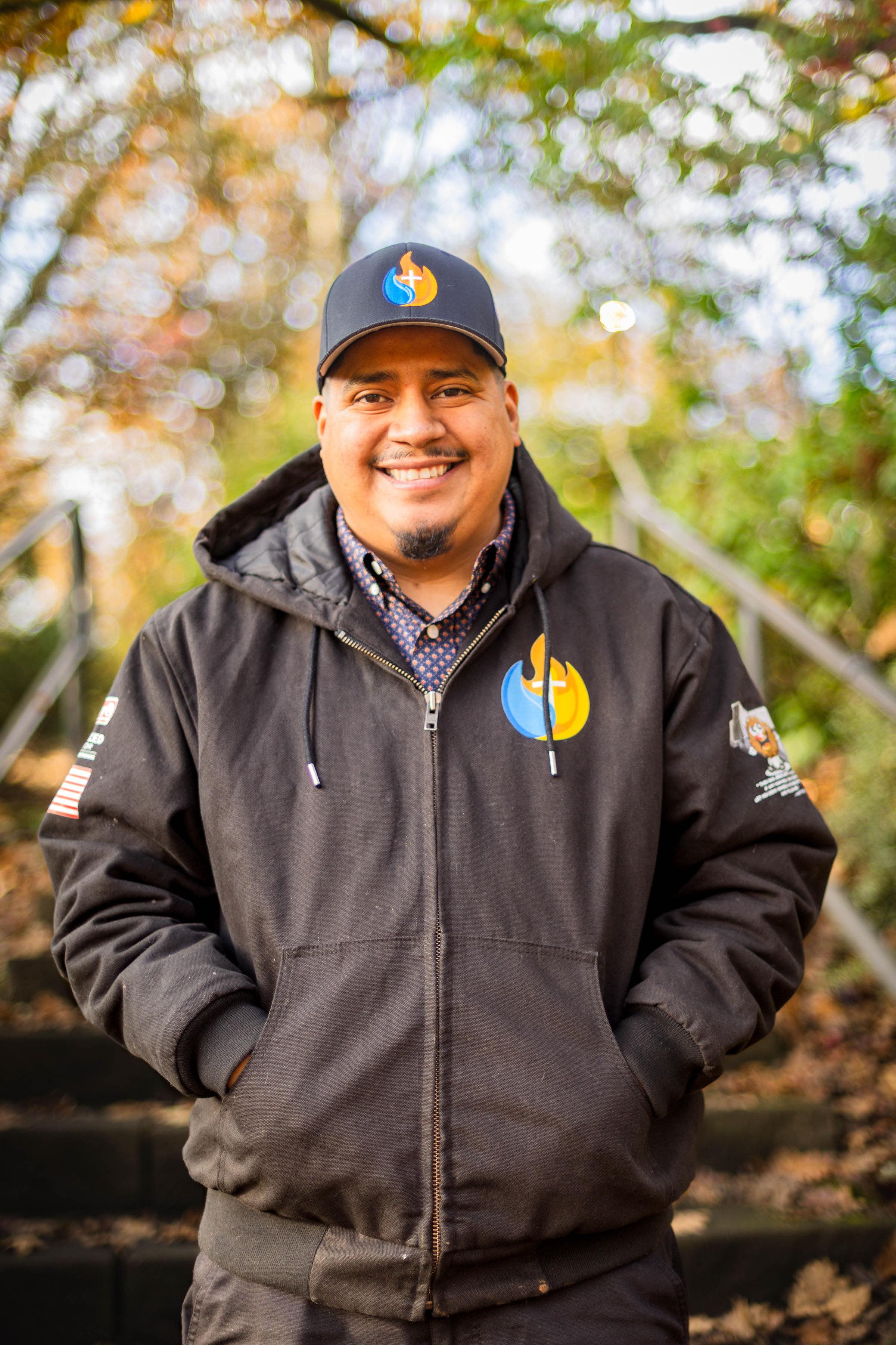 Eliseo Cortes, owner of HVAC Ministries, smiling in a branded company jacket and cap outdoors