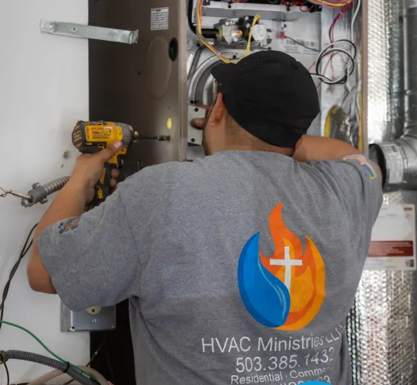 An HVAC Ministries technician in a branded shirt drilling into an open residential gas furnace during a repair